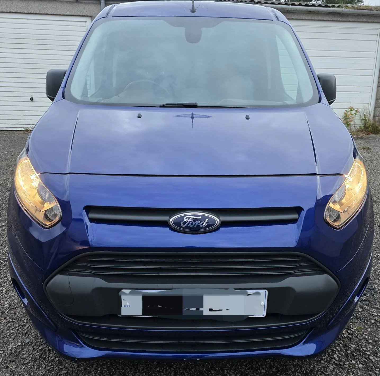 Front view of a blue Ford car with headlights on, parked in front of a white garage door.