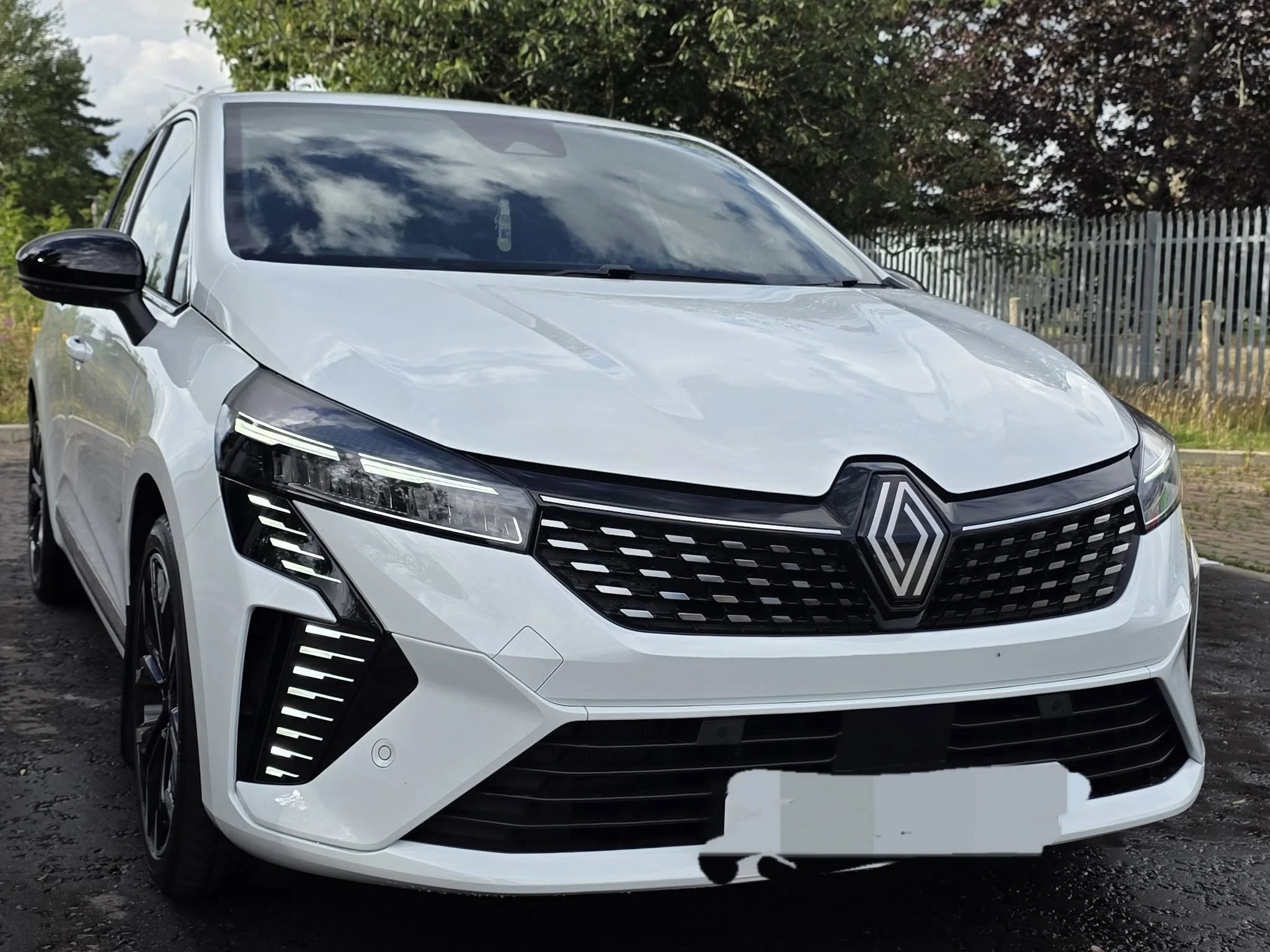 Front view of a white Renault car parked on a paved surface with trees and a fence in the background.