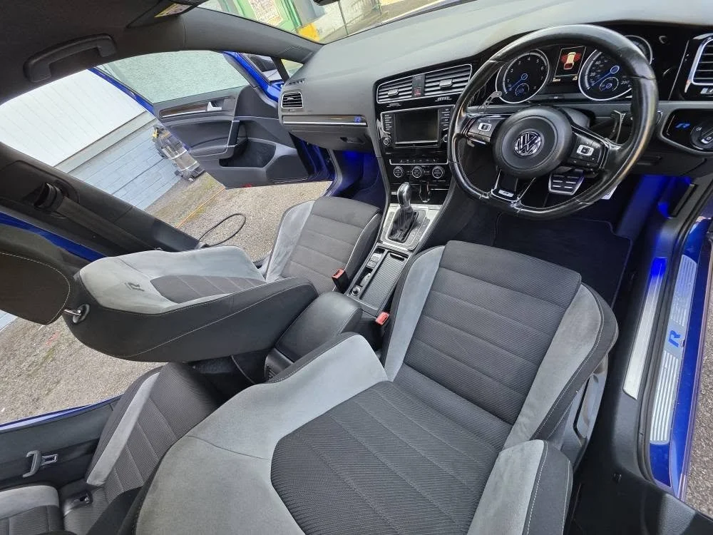 Interior of a Volkswagen vehicle, showing front and rear seats, steering wheel, dashboard with instrument cluster, center console with gear shifter, and an open driver-side door.