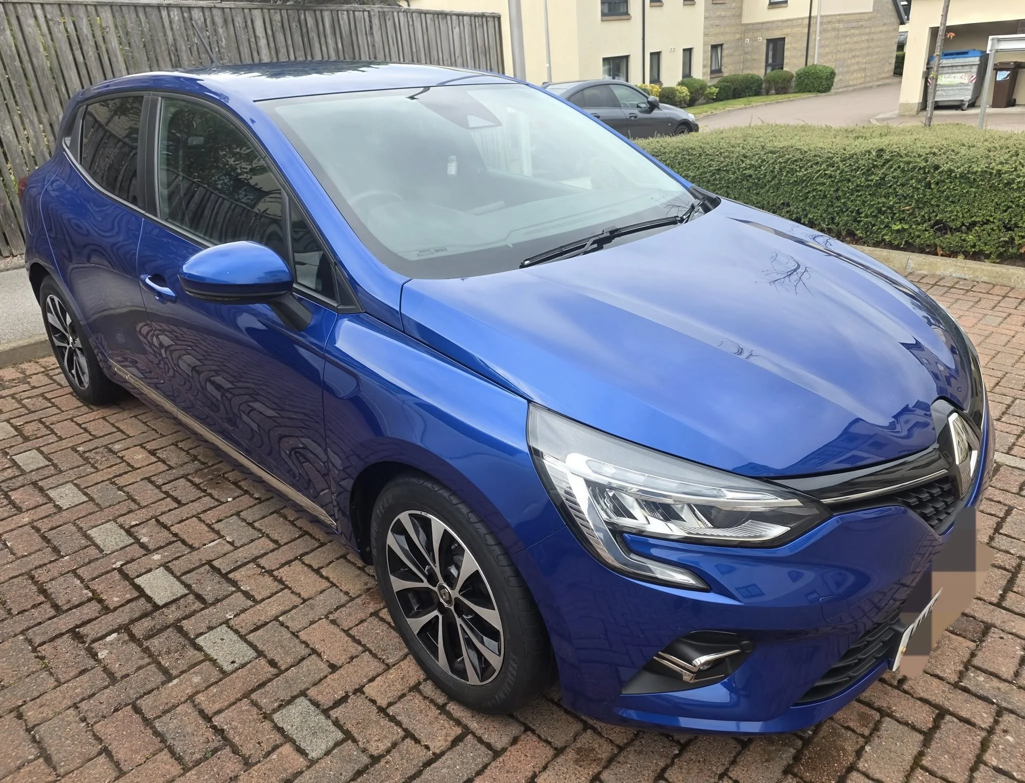 A blue hatchback car parked on a brick driveway in front of a wooden fence with residential buildings and greenery in the background.