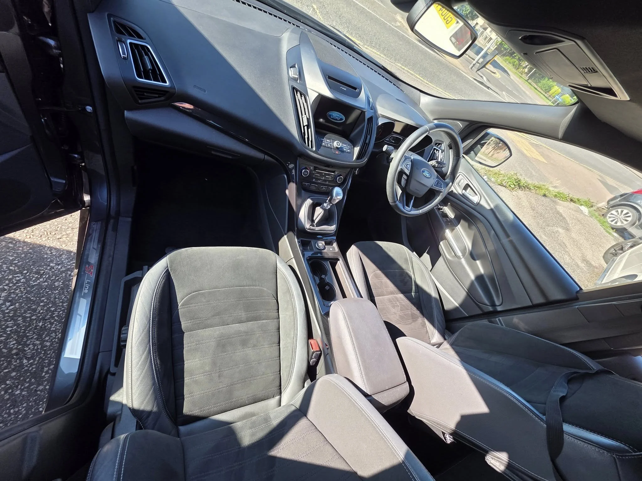 Interior of a Ford vehicle showing the dashboard, steering wheel, gear shift, front seats, and windshield with a view of parked cars outside.