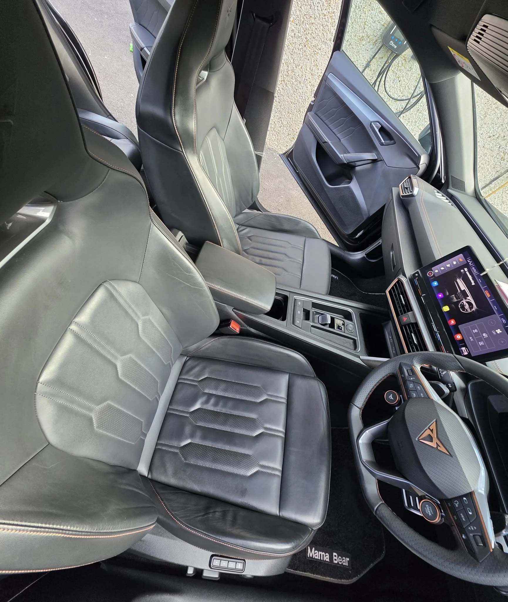 Interior view of a modern vehicle showing the driver and passenger seats, dashboard, steering wheel with a triangle logo, and a digital display on the center console. The seats are black with stitching, and the floor mat has the text 'Mama Bear'.