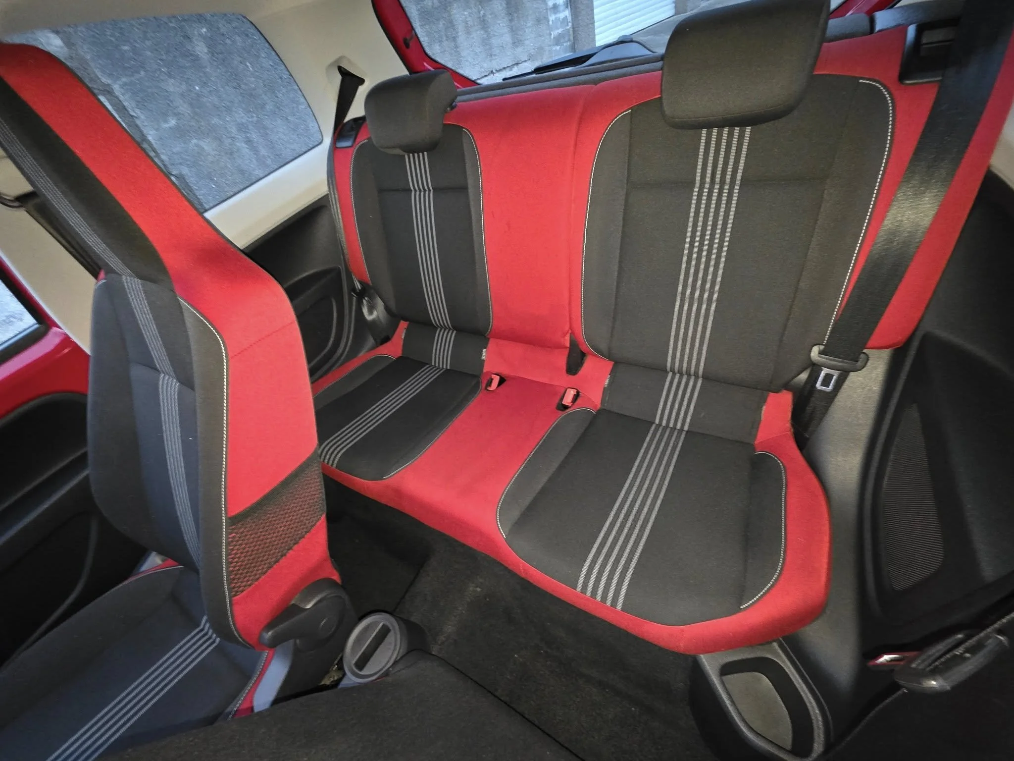 Red and black car rear seats with headrests and seat belts, inside a vehicle.