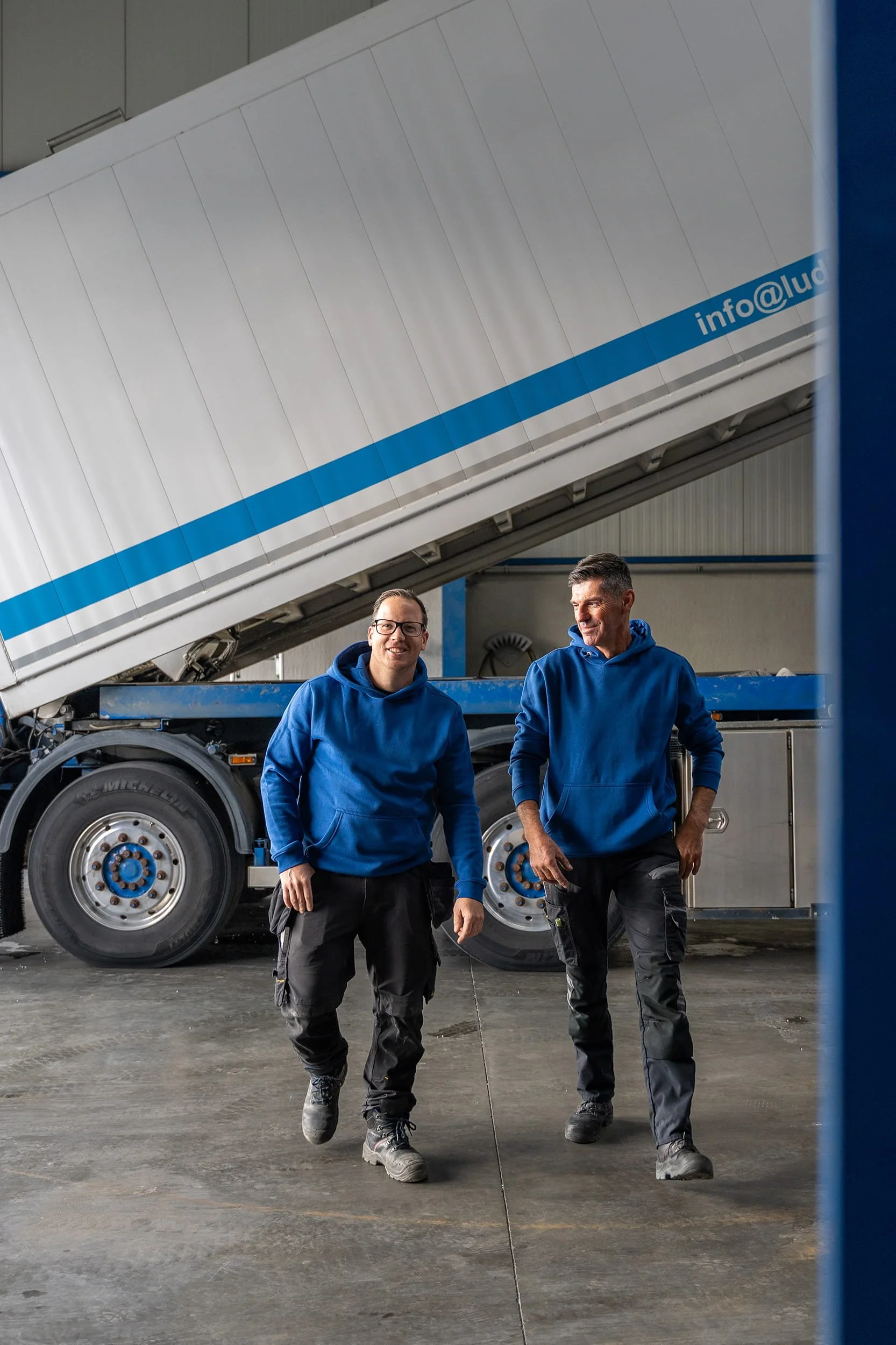 Twee medewerkers van Ludiso in blauwe hoodies lopen glimlachend weg van een vrachtwagen na het voltooien van het laadproces van isolatieproducten in de productiehal van Bilzen-Hoeselt.