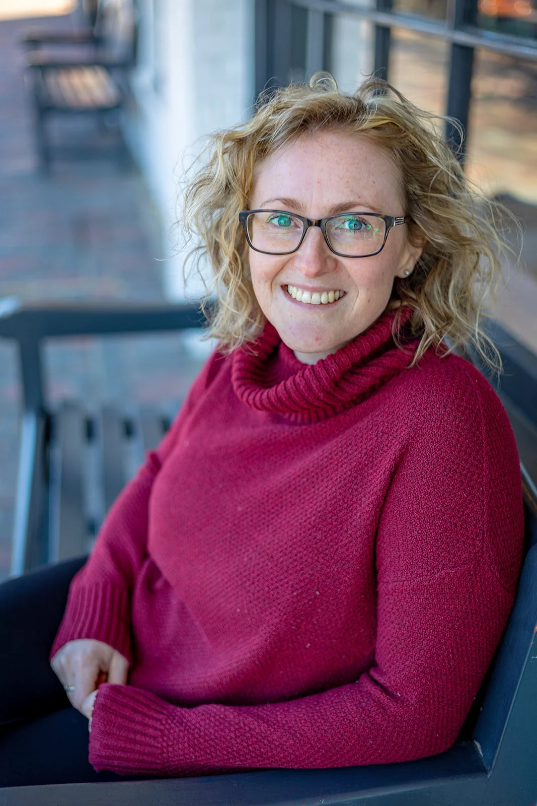 A woman with curly blonde hair and glasses wearing a red turtleneck sweater, sitting on a bench in front of a glass window, smiling at the camera.