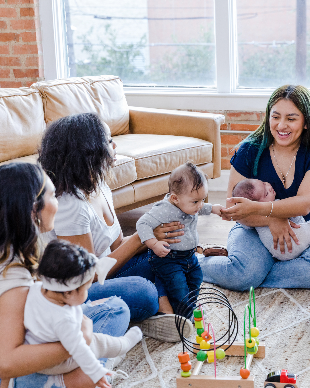 A group of women and children sitting on a carpeted floor in a living room, with one woman holding a newborn, while another child reaches out to the baby. There are colorful baby toys on the floor and a large window in the background.