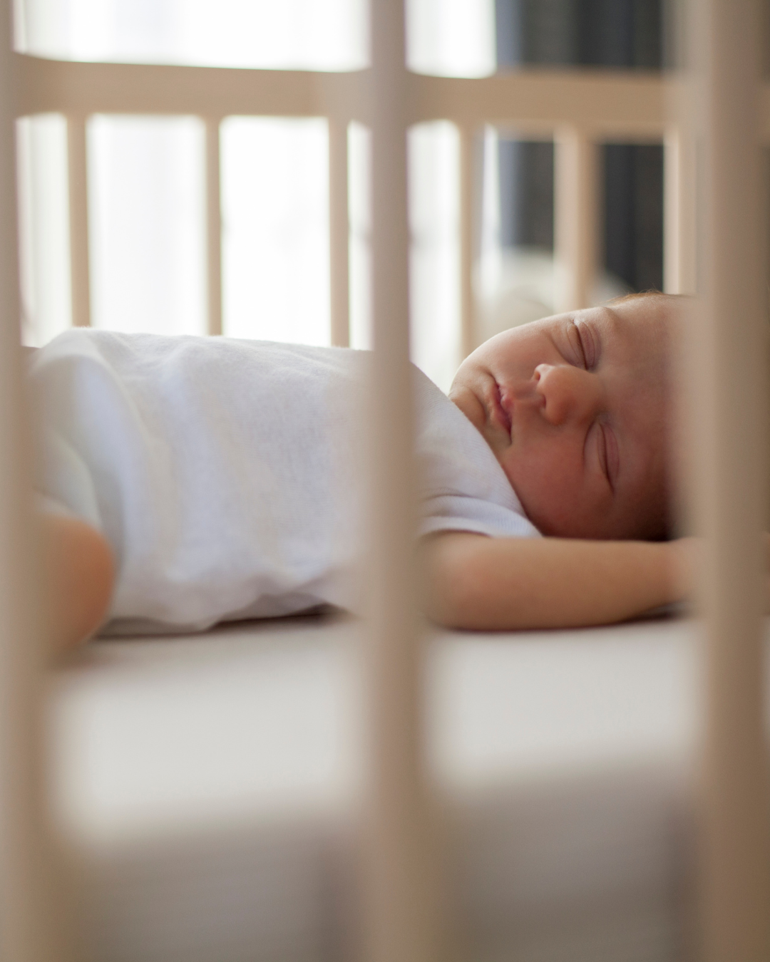 A sleeping baby lying on a crib mattress, seen through the crib bars.