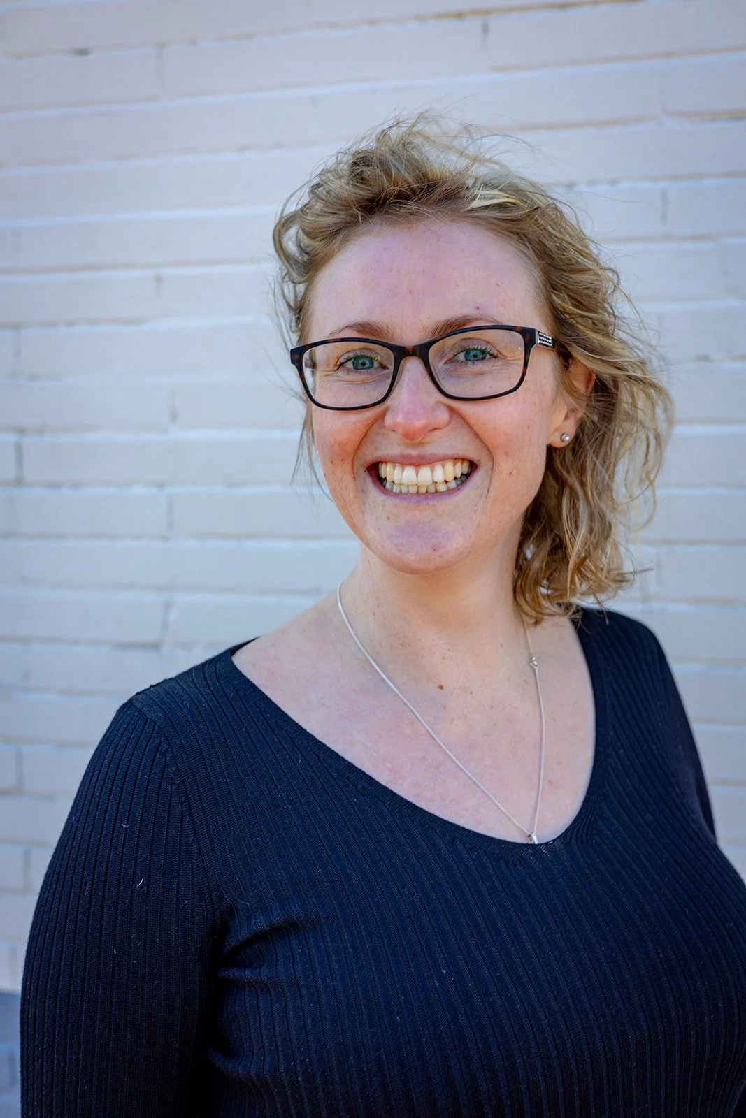A woman with glasses and curly blonde hair smiling in front of a white brick wall.
