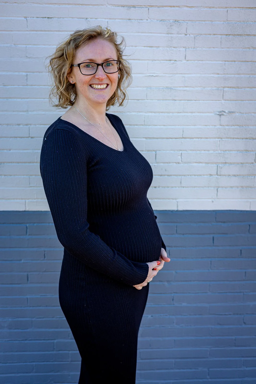 A woman with curly blonde hair, glasses, and a black ribbed sweater and pants standing in front of a white brick wall, smiling at the camera with her hands gently resting on her baby bump.