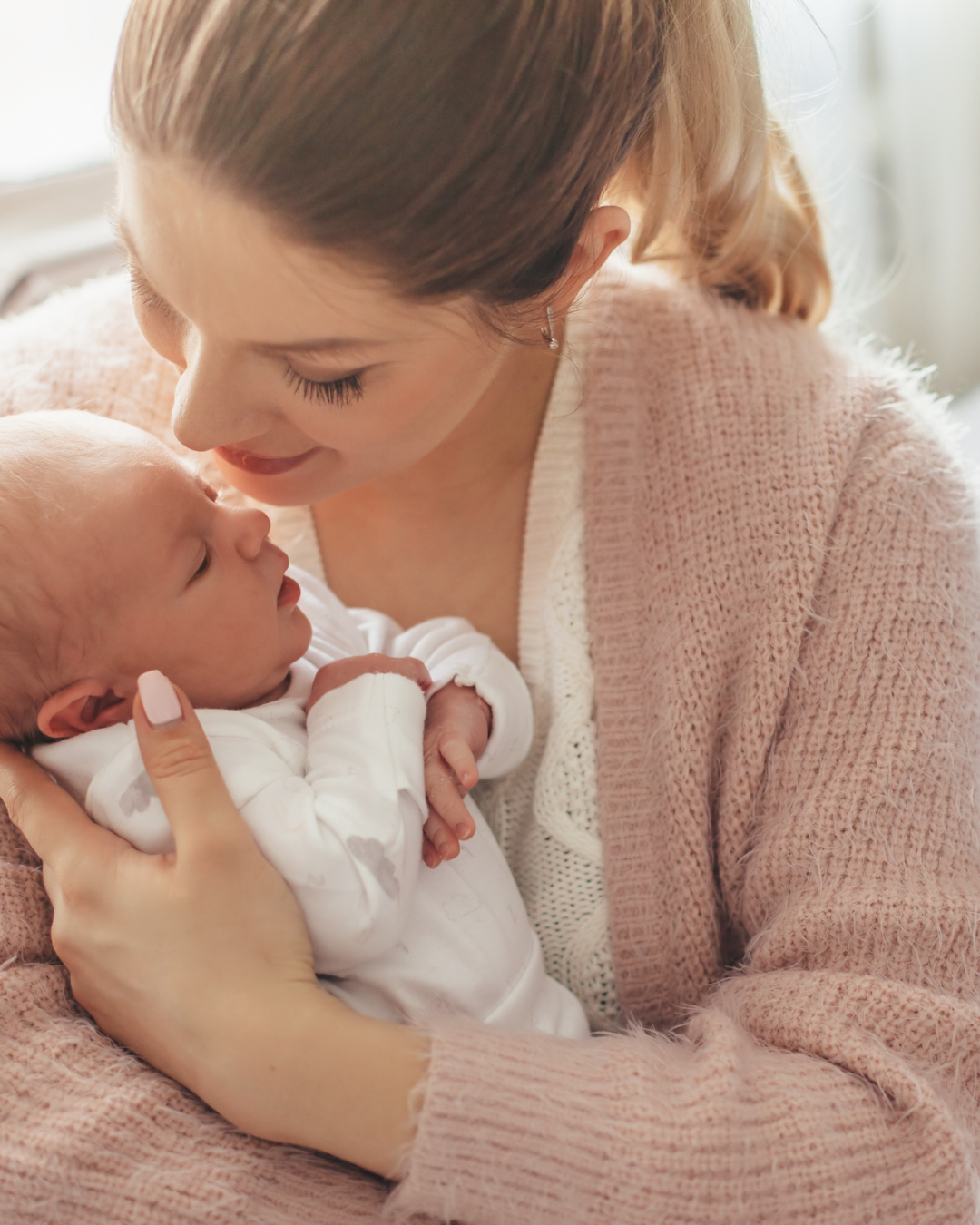 A woman gently holding a newborn baby close, both looking at each other lovingly indoors.