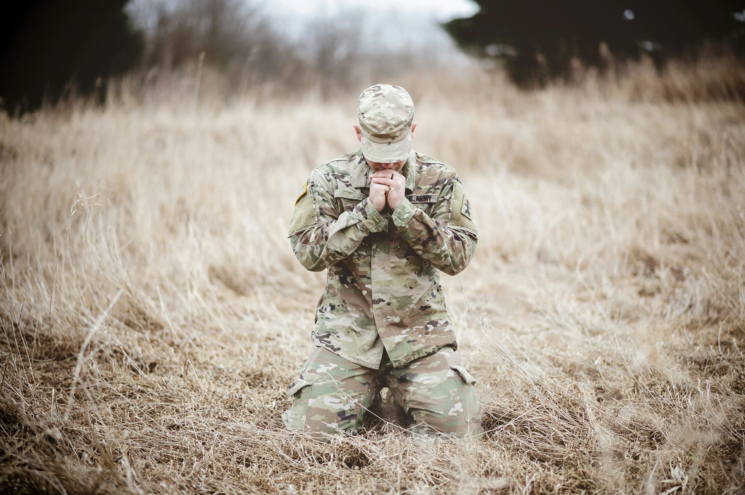 A soldier in camouflage uniform kneels in dry, beige grass with hands clasped in prayer, head bowed, in an open field with blurred trees in the background.