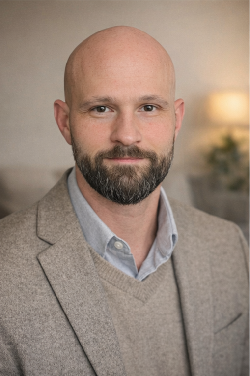 Portrait of a bald man with a beard wearing a light gray blazer over a blue shirt, standing indoors with soft lighting and blurred background.