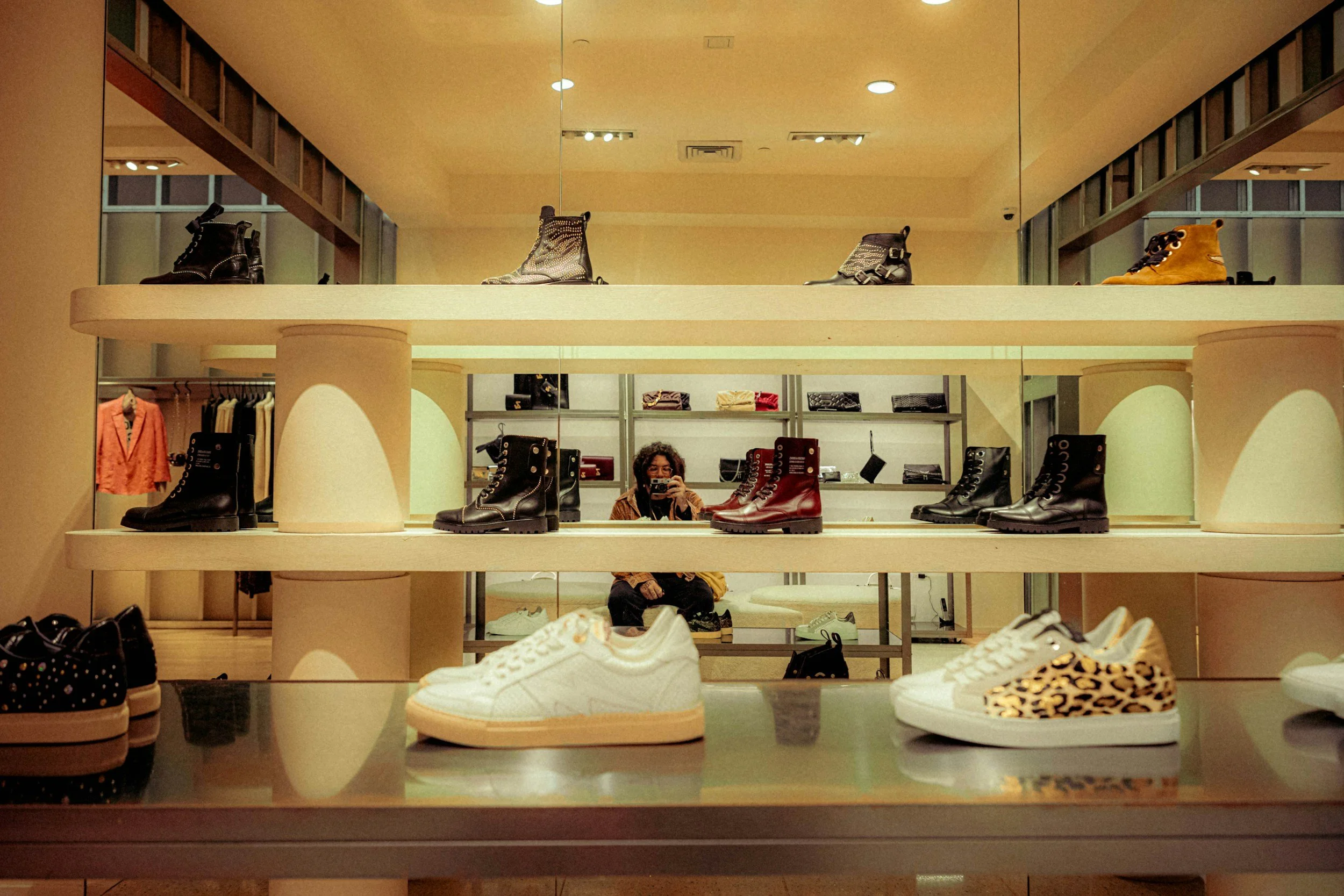 Display of various shoes including boots, sneakers, and leopard print shoes in a retail store, with a person taking a photo in the mirror reflection.