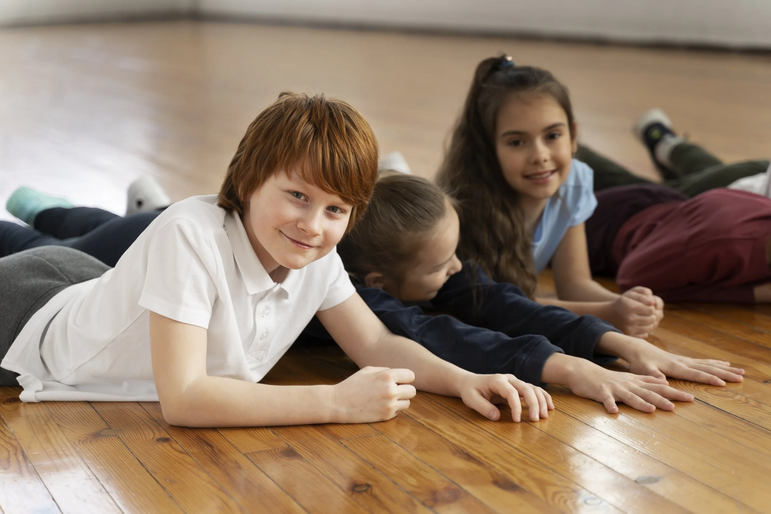 Three children lying on a wooden floor, smiling and stretching, in a casual indoor setting.