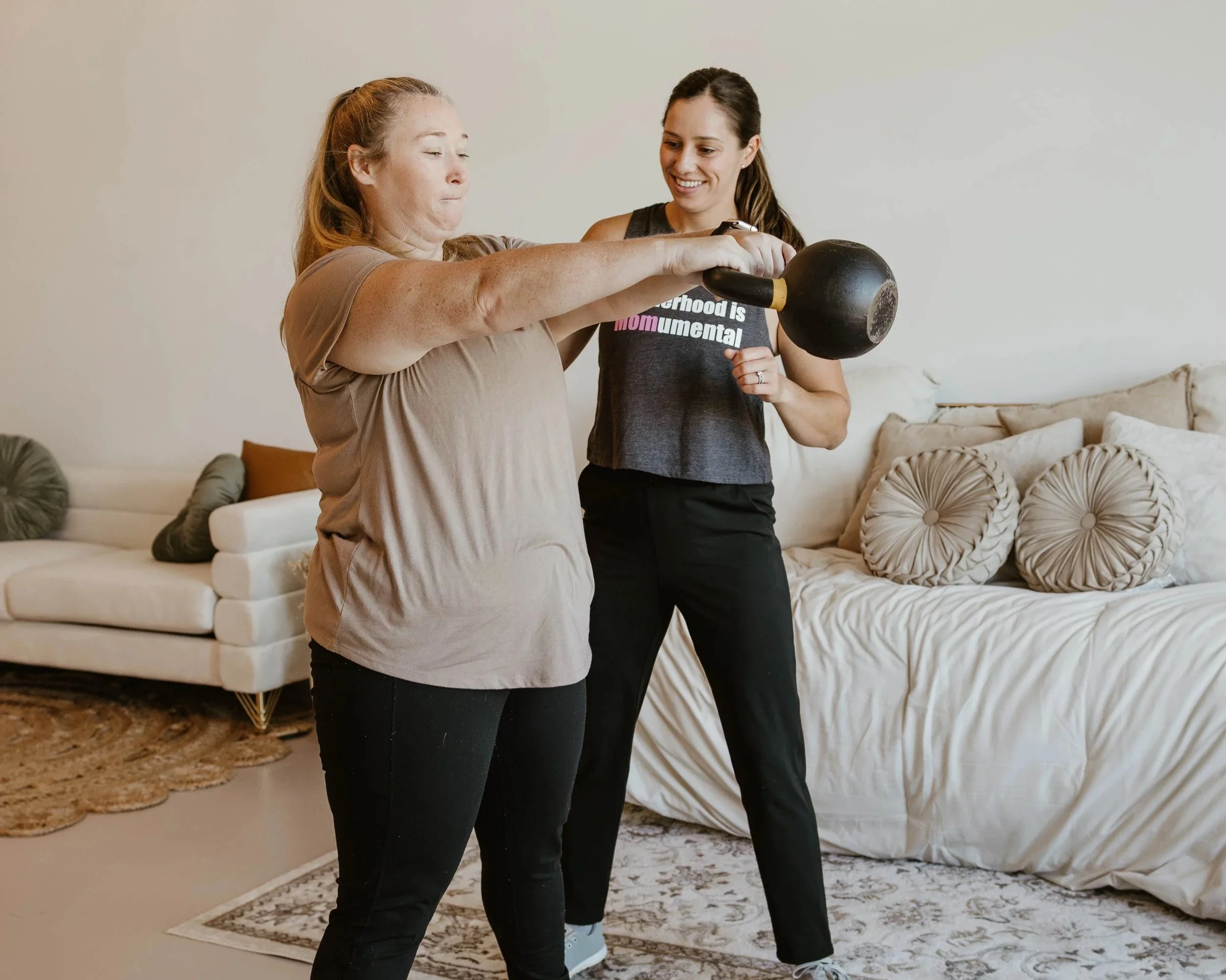 Two women inside a living room, one helping the other hold a kettlebell for exercise, with a beige sofa and decorative pillows in the background. Pelvic floor physical therapy, women's health physical therapy, orthopedic physical therapy.