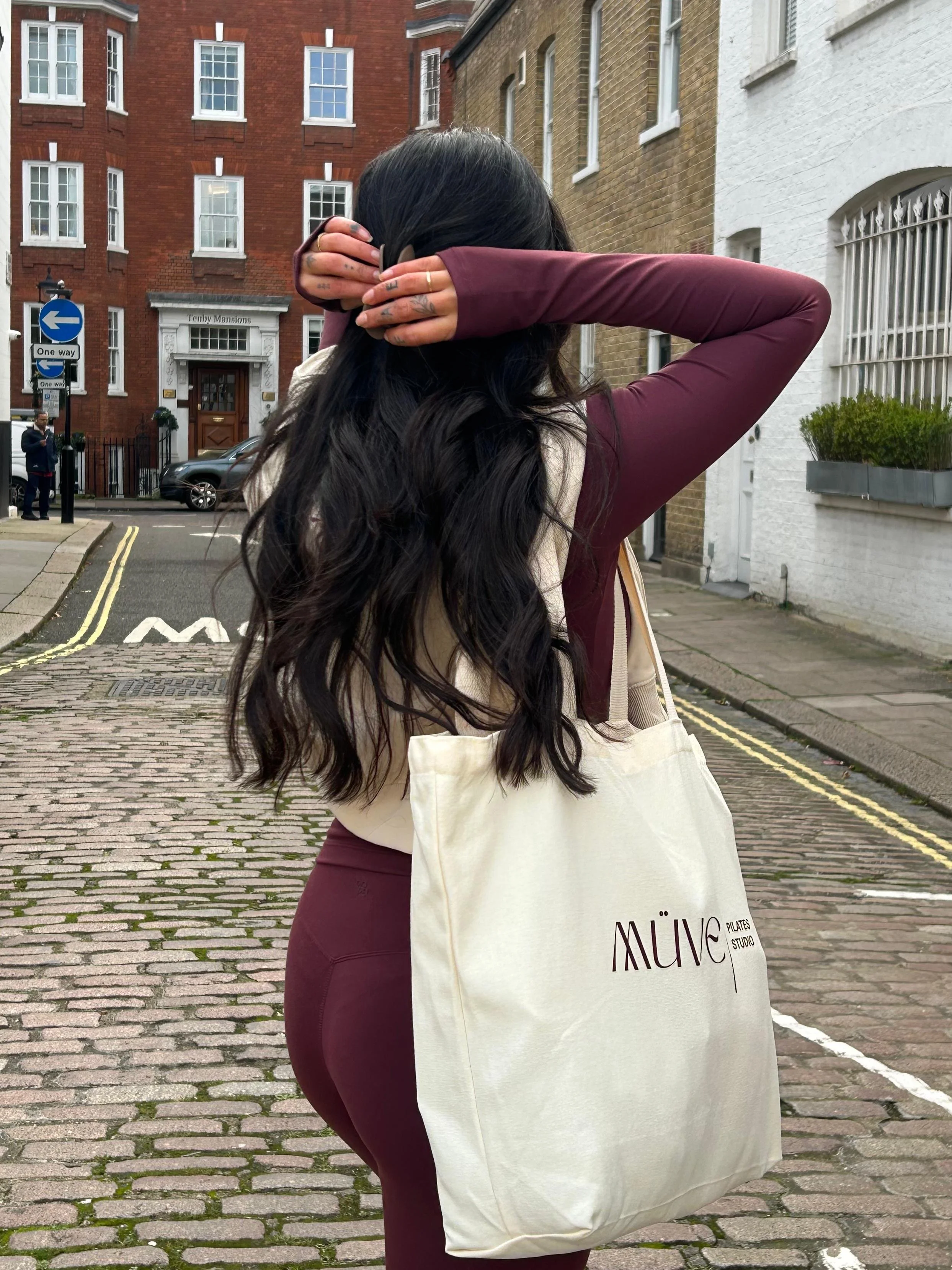 A woman with long black hair, wearing a beige top and maroon leggings, is standing on a cobblestone street, holding her hair back with her arms behind her head. She is carrying a large beige tote bag with the words 'MÜVE PLATES STUDIO' printed on it.