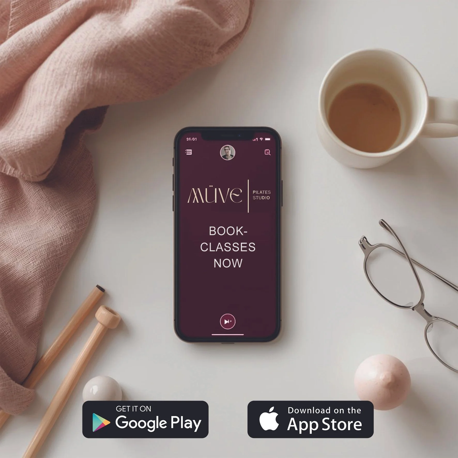 Smartphone on white table displaying the ALMAVE Pilates Studio app with booking options, surrounded by a pink cloth, a cup of coffee, reading glasses, and decorative items.