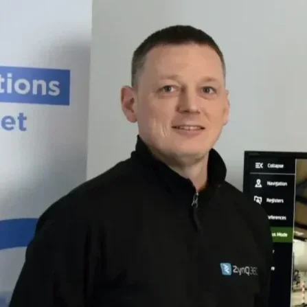 A man with short brown hair, wearing a black zip-up jacket with a logo, standing in front of a display screen and a banner with blue text.