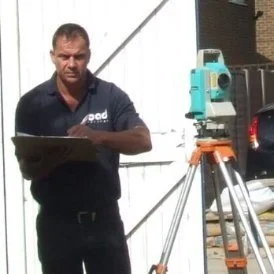 Man standing outdoors in front of a white fence, operating a theodolite mounted on a tripod.