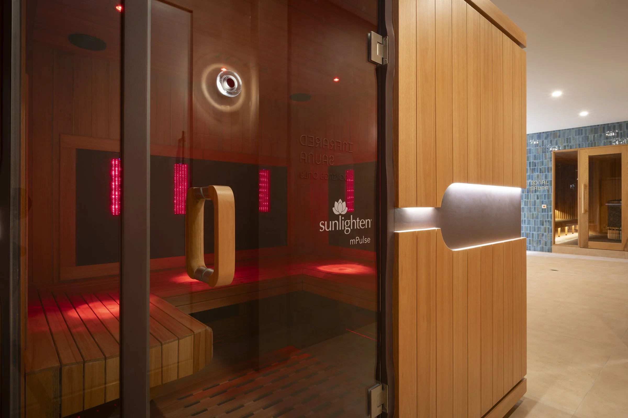 Indoor sauna room with glass door, wooden interior, red infrared lights, and a wooden bench, located in a wellness facility with seating area and blue tiled wall in the background.