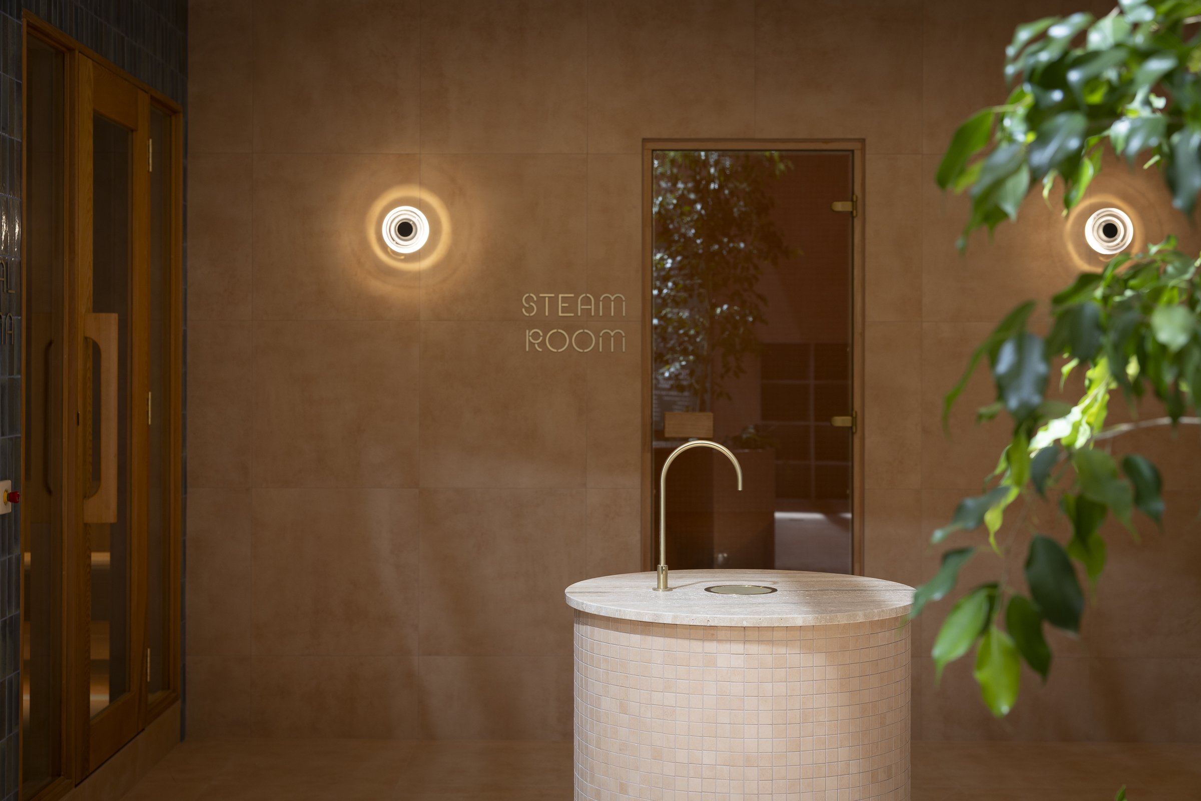 A steam room with warm lighting, a small round tiled fountain with a faucet, a glass door, and wall-mounted lights, along with a green plant partially visible on the right.
