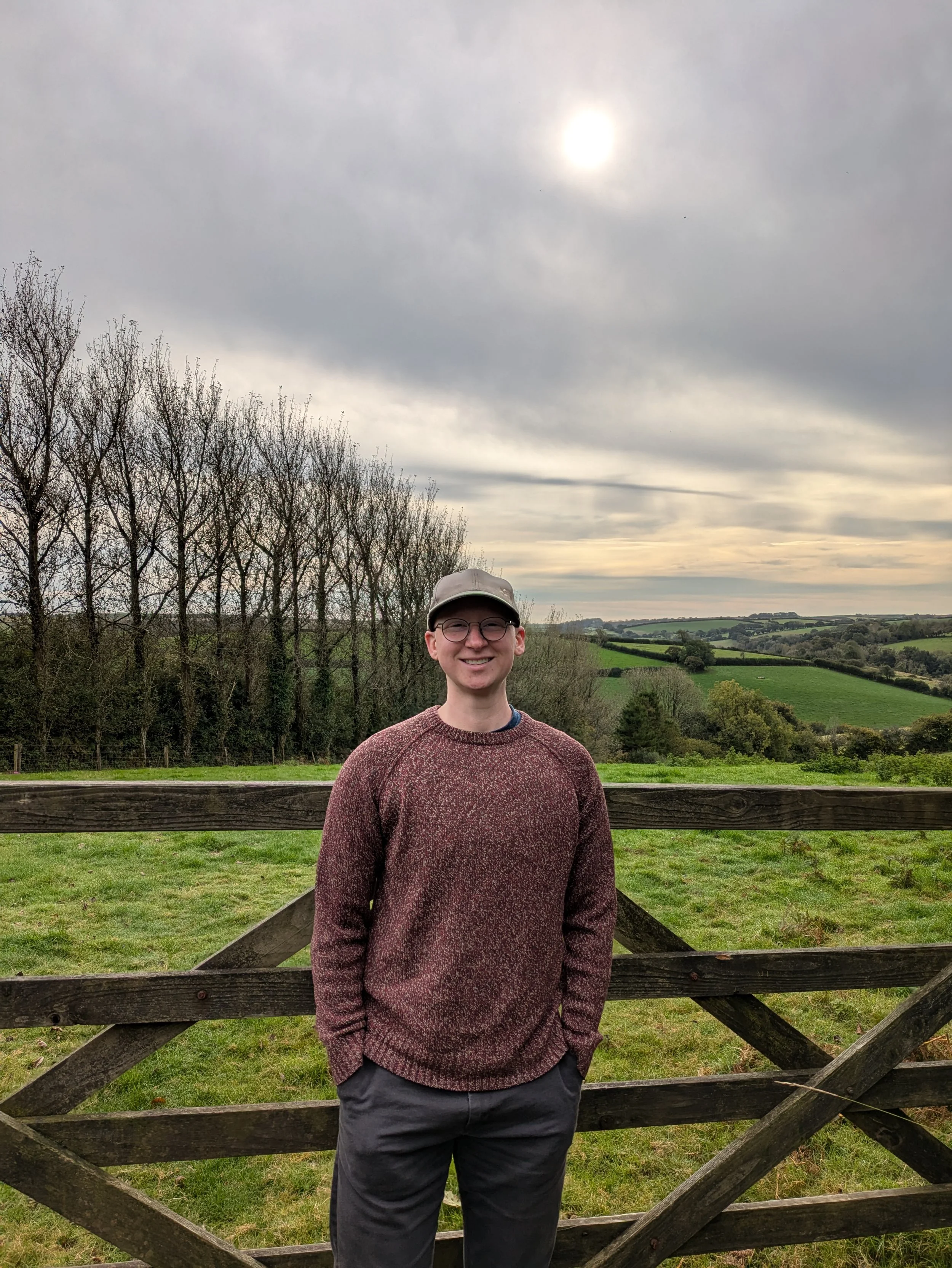 A smiling person wearing glasses, a beige cap, a maroon sweater, and gray pants standing outdoors in front of a wooden fence, with a landscape of green fields, trees, and cloudy sky in the background.