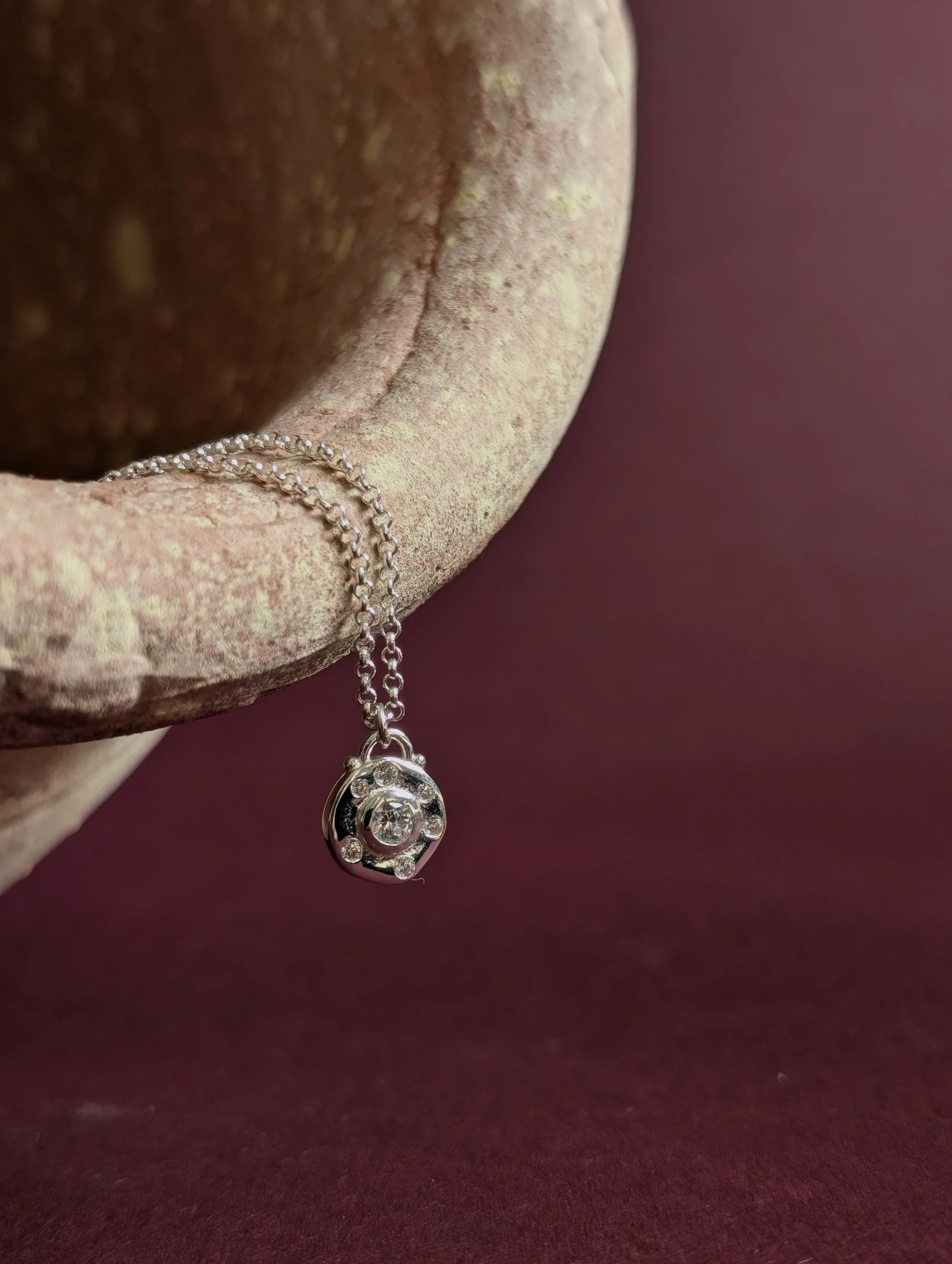 A silver necklace with a circular pendant featuring a central gemstone and surrounding smaller gemstones, hanging from a rough, pinkish stone surface against a dark mauve background.
