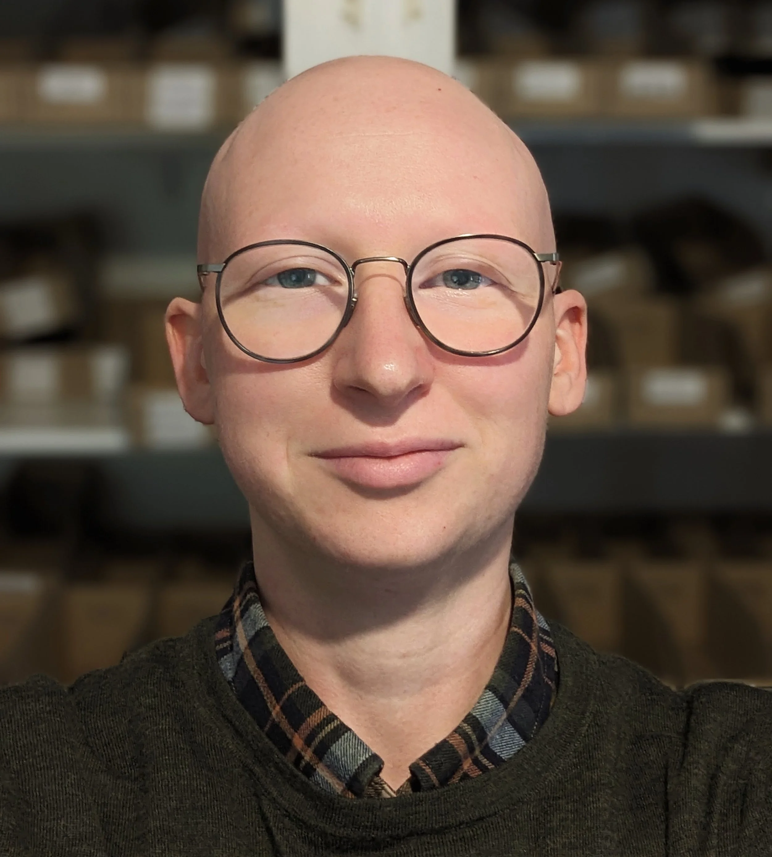 A person with a bald head, round glasses, and fair skin, smiling in front of shelves filled with boxes.