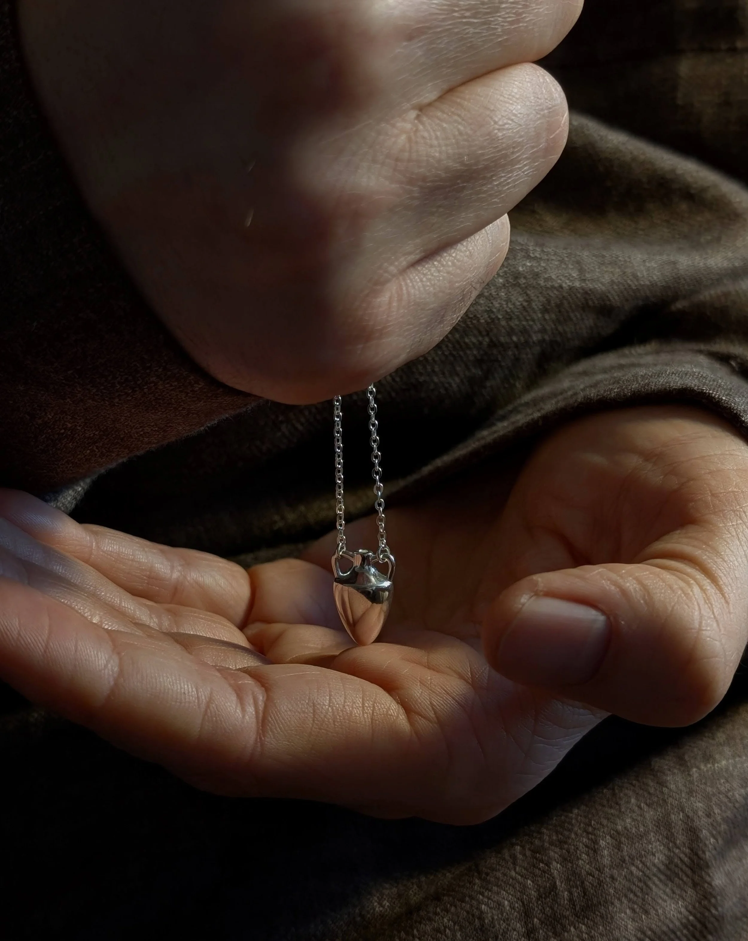 A close-up of a person's hand holding a silver necklace with a small, shiny, metallic pendant shaped like a heart.