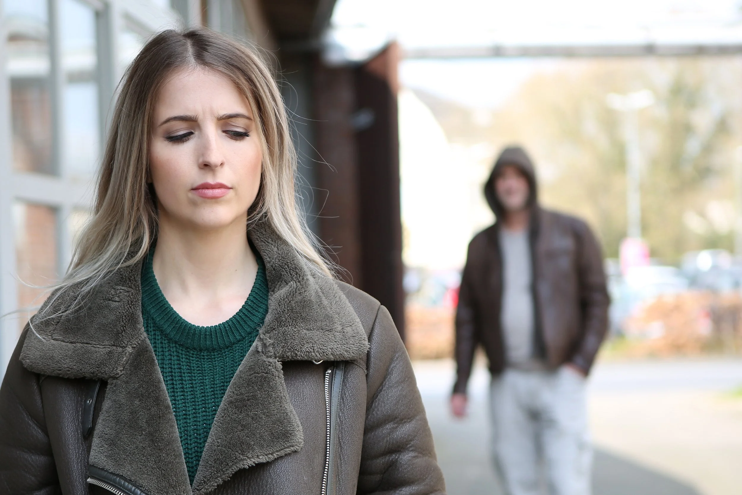A young woman with blonde hair and a serious expression stands outside with her eyes closed, wearing a brown jacket over a green sweater. In the background, a man wearing a hooded jacket stands blurred in an outdoor setting.