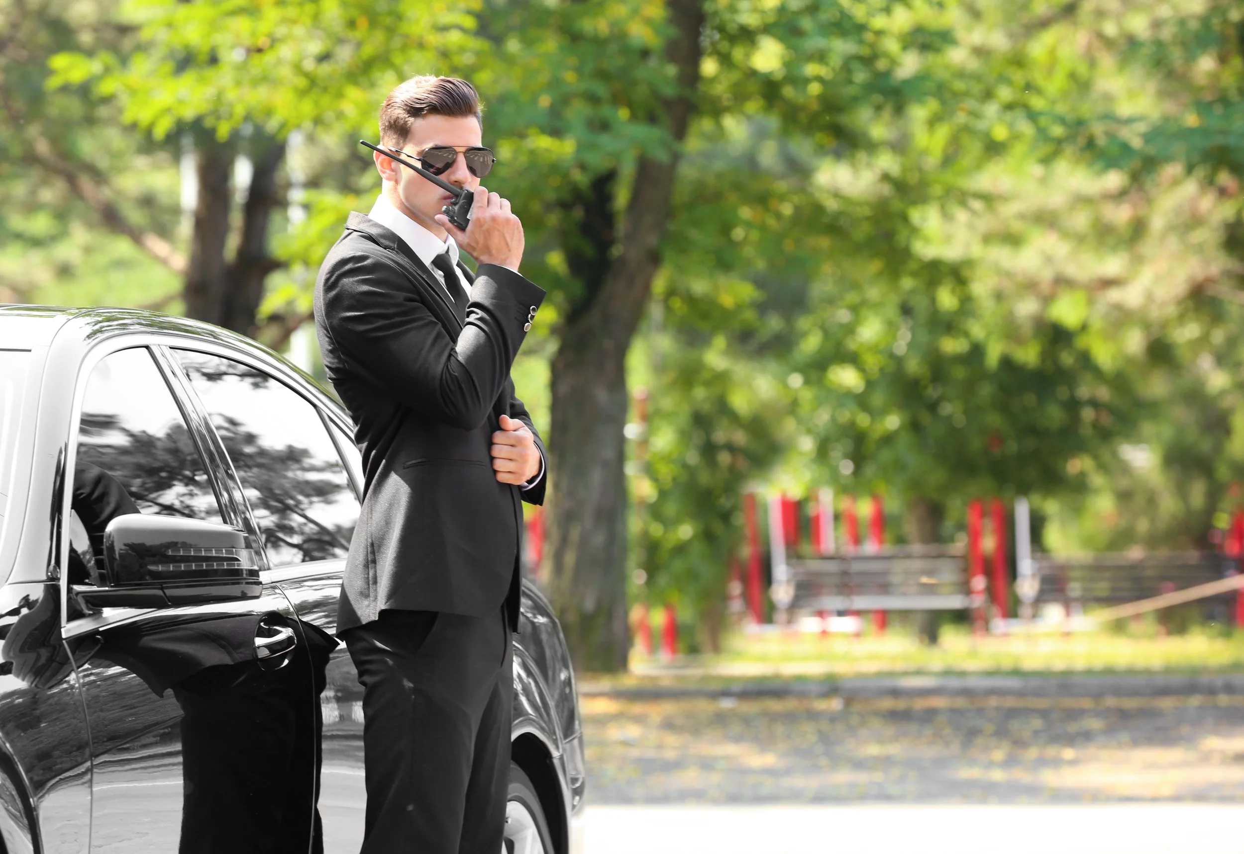 Man in black suit and sunglasses holding a walkie-talkie standing next to a black car outdoors with trees in the background.