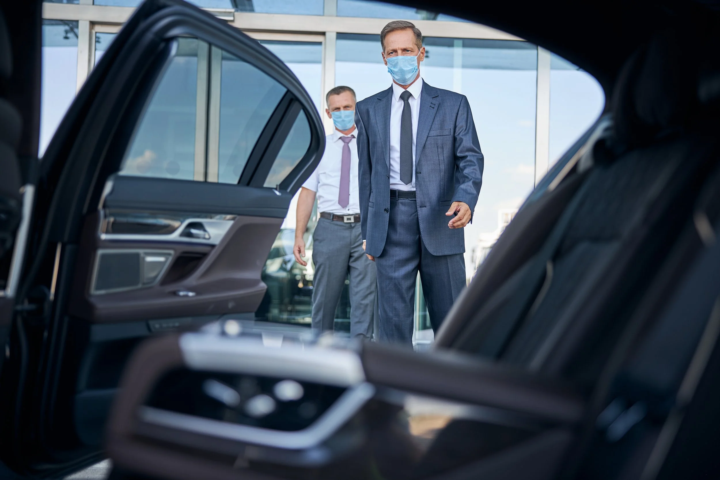Two men in suits and face masks standing outside near a car, viewed from the inside of the vehicle.