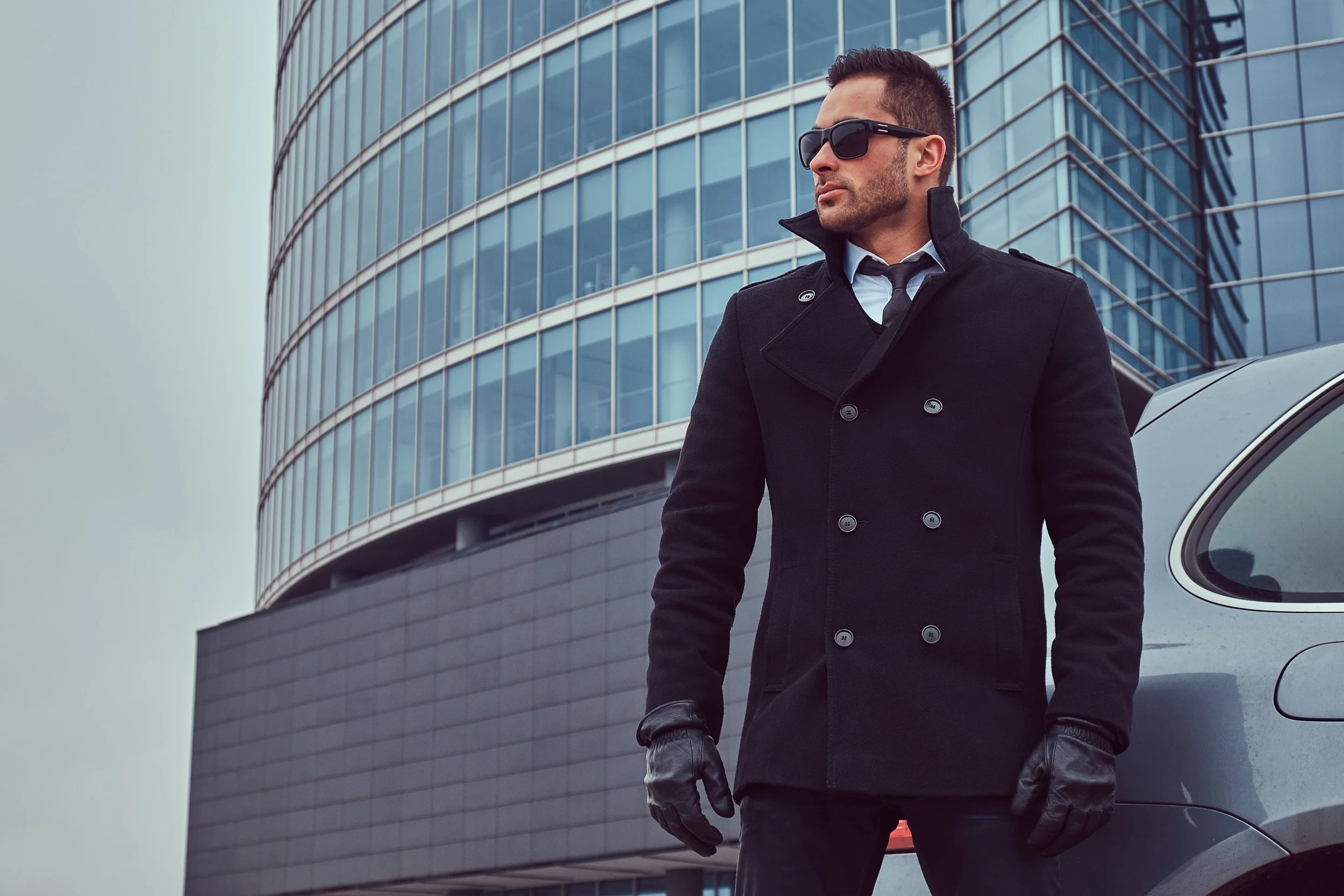 Professional bodyguard wearing a dark coat and sunglasses standing outside a modern office building, symbolizing Fortitude Security’s expert security team in Essex.