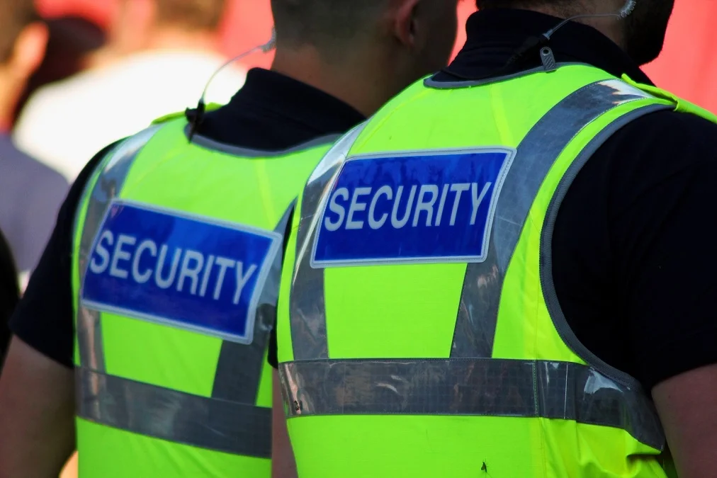Two security personnel wearing high-visibility yellow vests with blue 'SECURITY' patches on the back, standing outdoors.