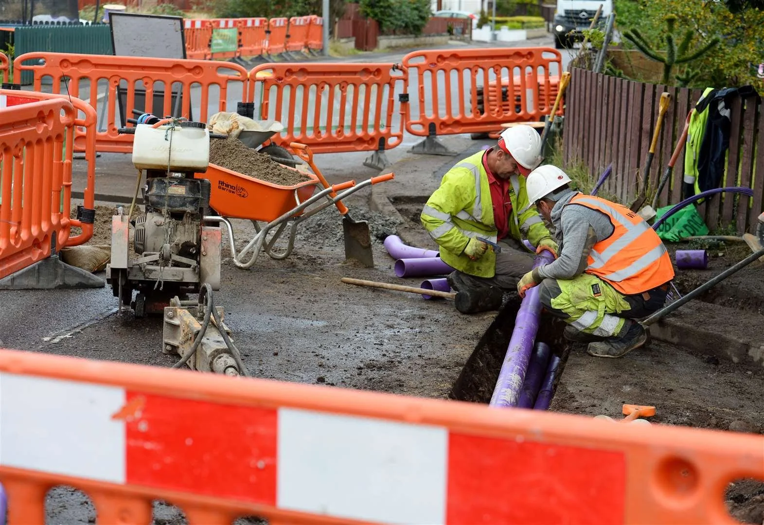 Construction workers in safety gear working on underground pipeline installation on a city street, with orange barriers and construction equipment around.