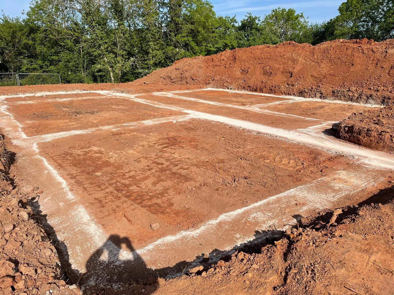 A construction site with marked rectangular areas on the ground for foundation footings, surrounded by dirt and soil with trees in the background.