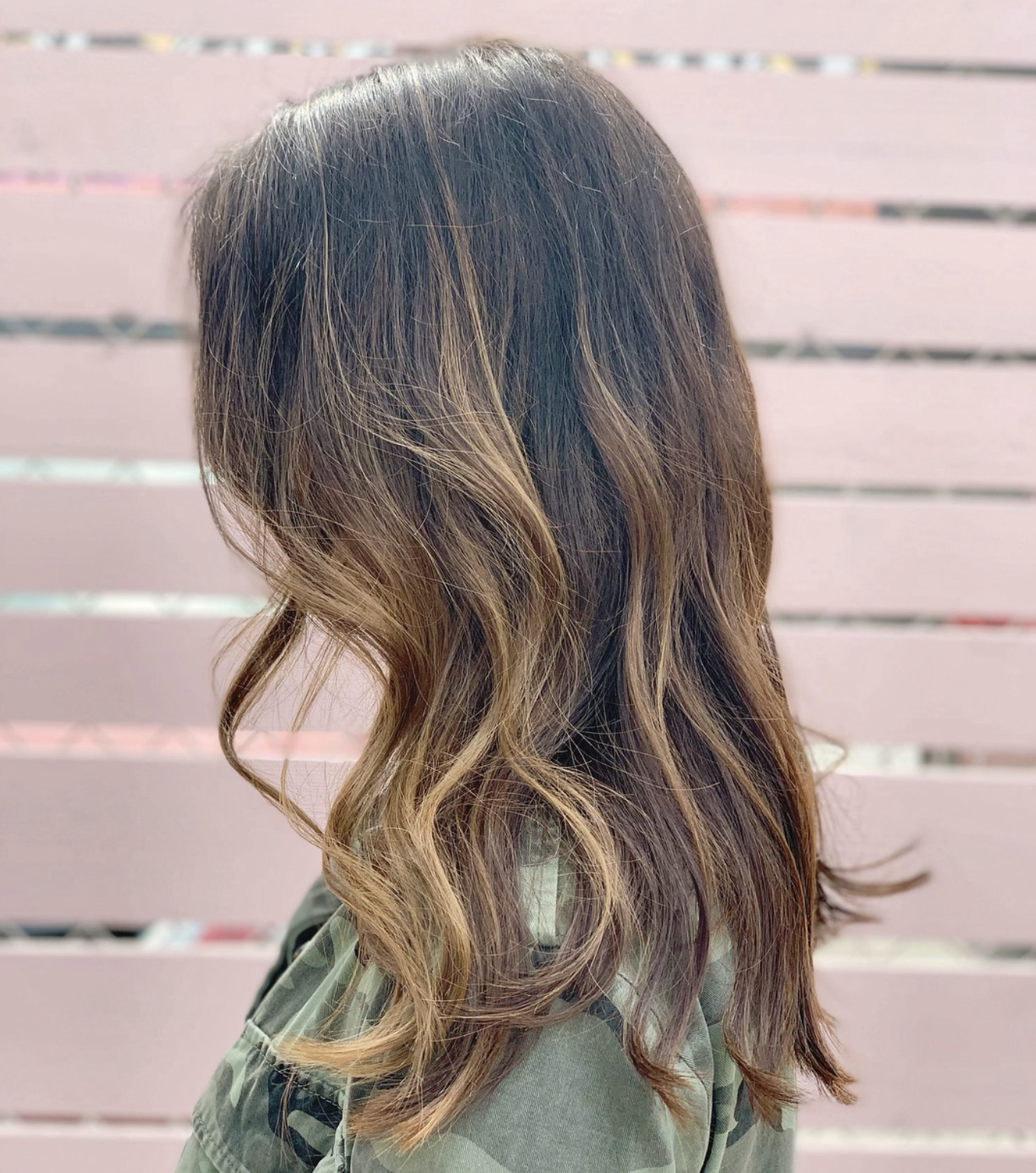 Side view of a woman with wavy, ombre hair in shades of brown and blonde, wearing a camouflage jacket, standing in front of a pink wooden fence.