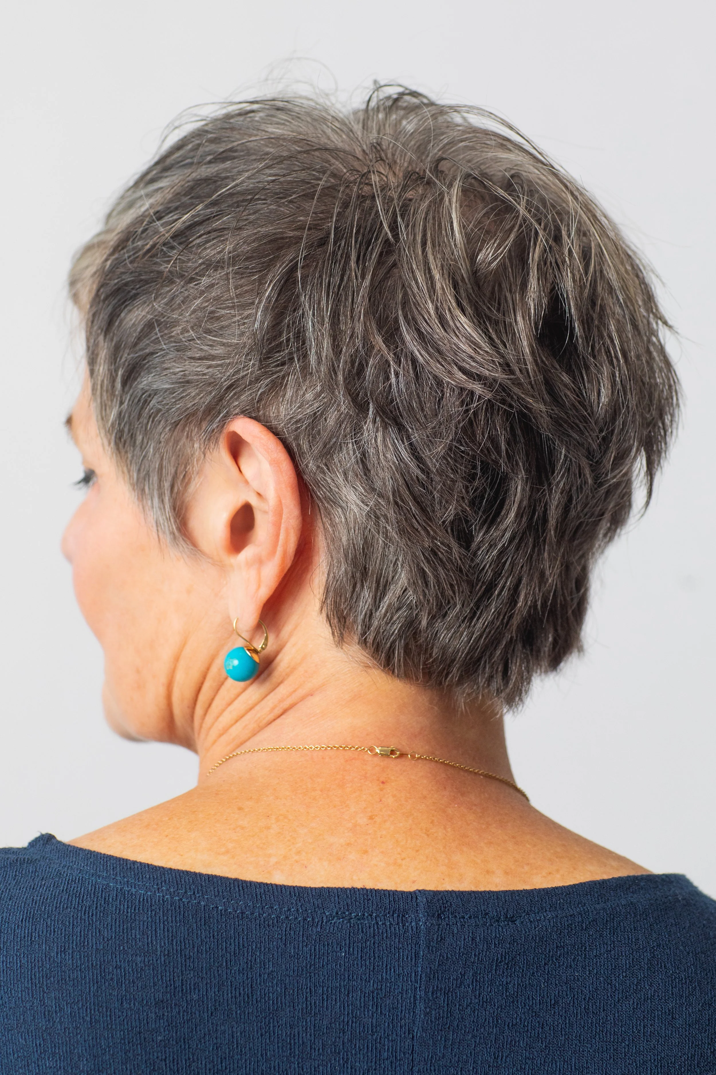 Back view of a textured short cut that celebrates natural gray growth. Layering and shaping create softness and lift, making this an ideal style for clients seeking easy wash-and-go hair. gray hair blending, short layered haircut, wash-and-go hair At