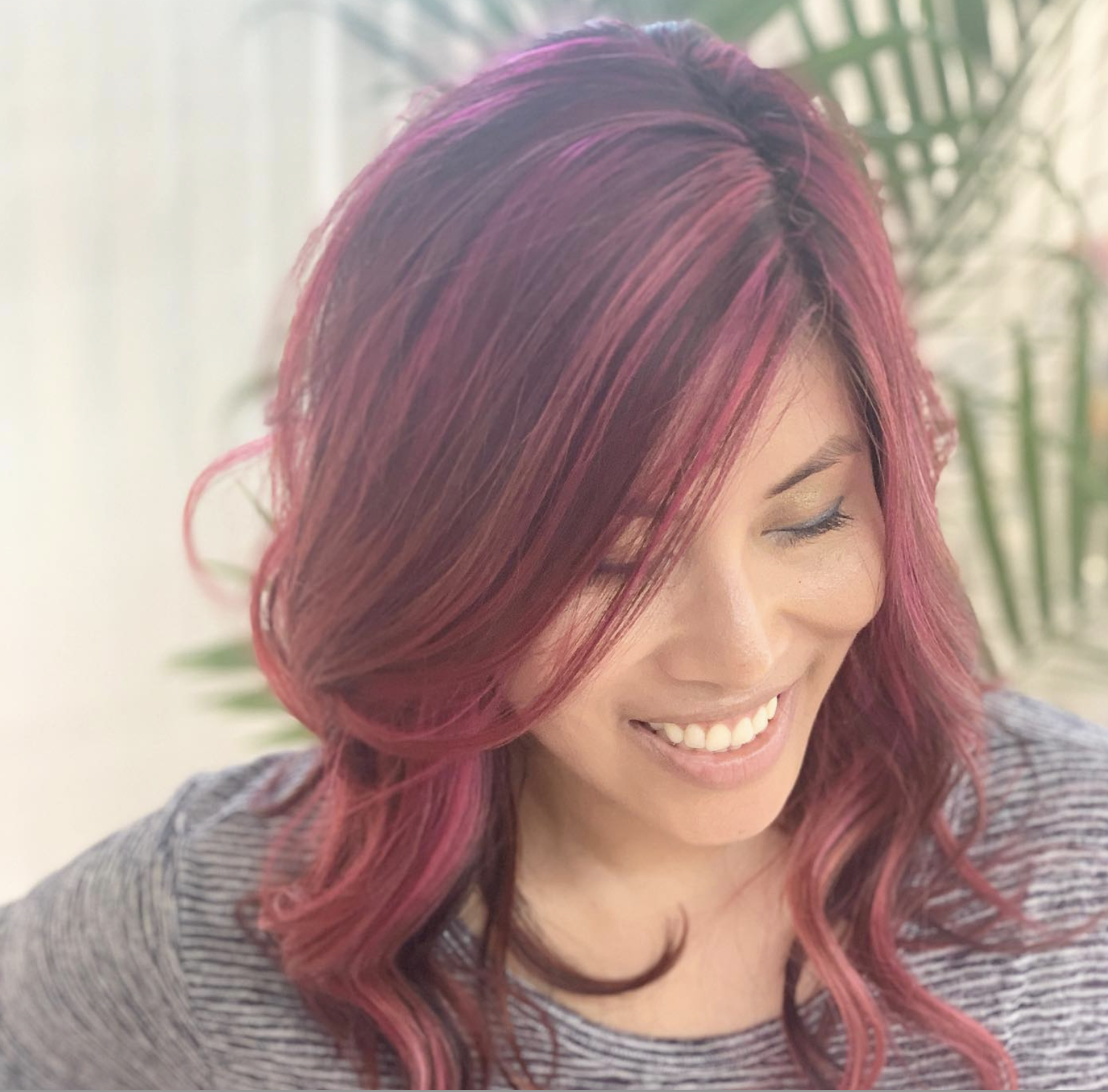 Woman with wavy, shoulder-length pink and purple hair, smiling and looking downward, in front of a blurred background of green plants.