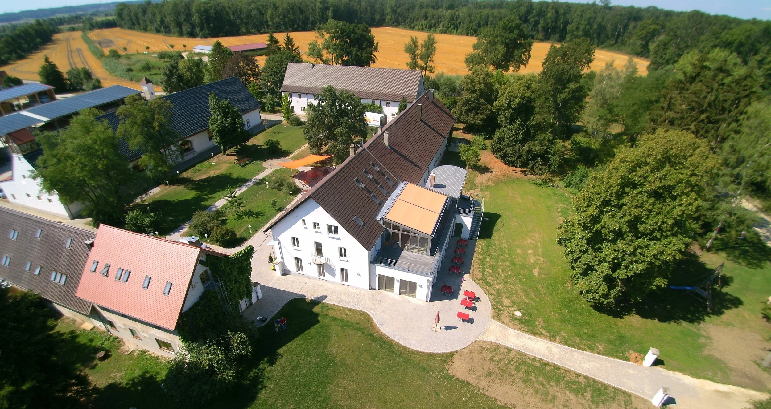 Luftaufnahme eines weißen Hauses mit rotem Dach, umgeben von grünem Garten und Bäumen, im Hintergrund Felder und Wald.