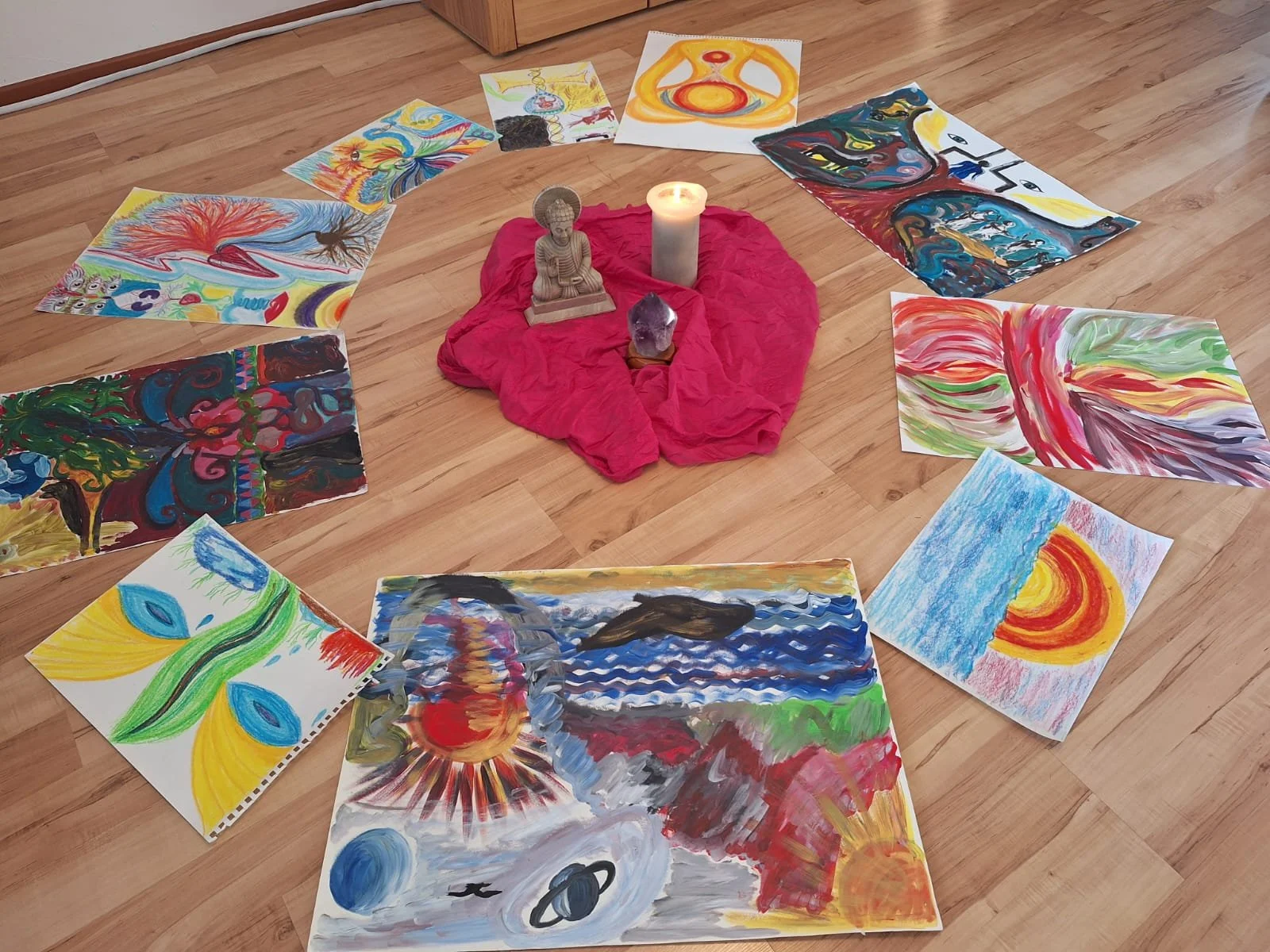 Kunstwerke und Zeichnungen in bunten, abstrakten und realistischen Stilen sind kreisförmig um eine kleine Sitzgelegenheit mit einer Skulptur, einem Kerzenlicht und einem pinken Tuch auf dem Holzboden angeordnet.