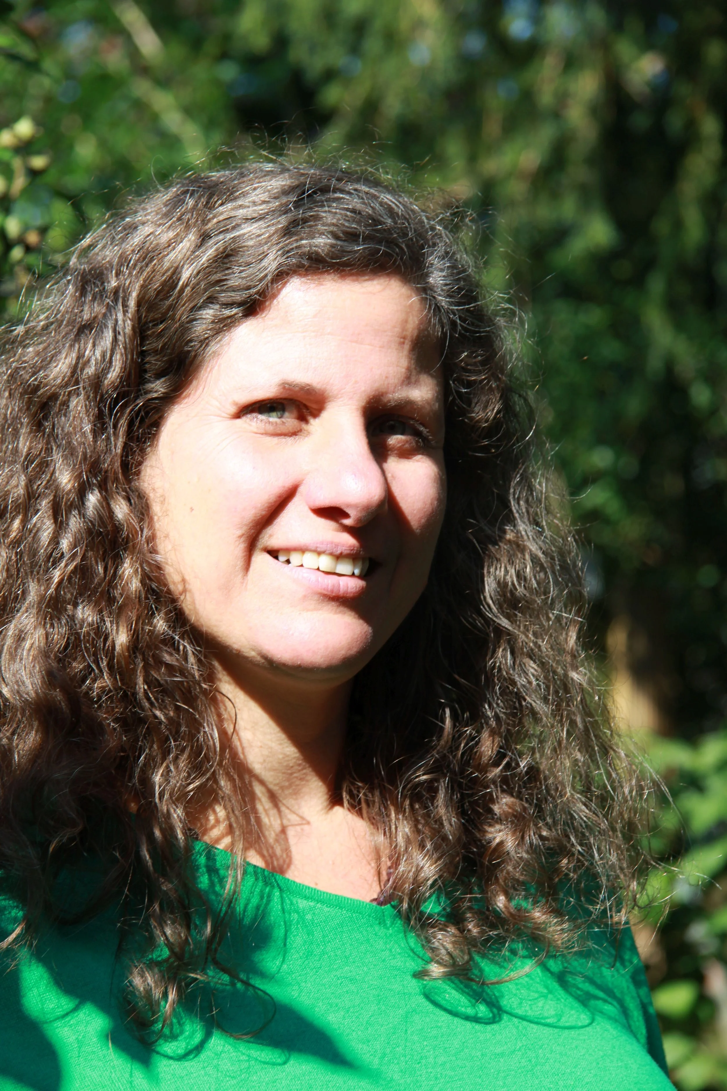 Nahaufnahme von Sandra Mahr mit langen, lockigen braunen Haaren, die Sonnenlicht auf ihr Gesicht wirft, im Freien vor einem grünen, bewaldeten Hintergrund, trägt grünes Shirt.