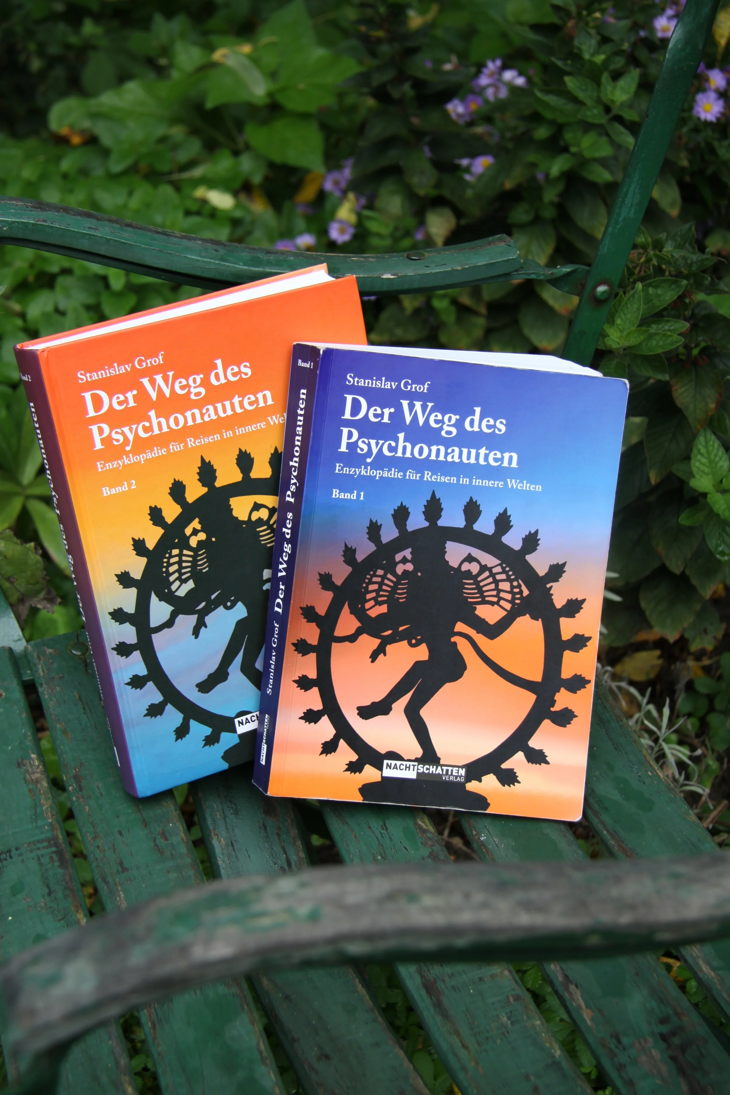 Zwei Bücher mit dem Titel 'Der Weg des Psychonauten' auf einer grünen Gartenbank, umgeben von Pflanzen. Das eine Buch ist orange und das andere blau, beide zeigen ein mystisches Symbol auf dem Cover.
