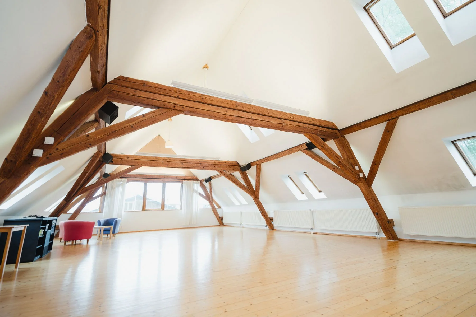 Ein heller, großer Raum mit Holzfußboden und sichtbaren Holzdeckenbalken. Mehrere Dachfenster lassen natürliches Licht strömen. Einige kleine Möbelstücke sind in der Ecke sichtbar.