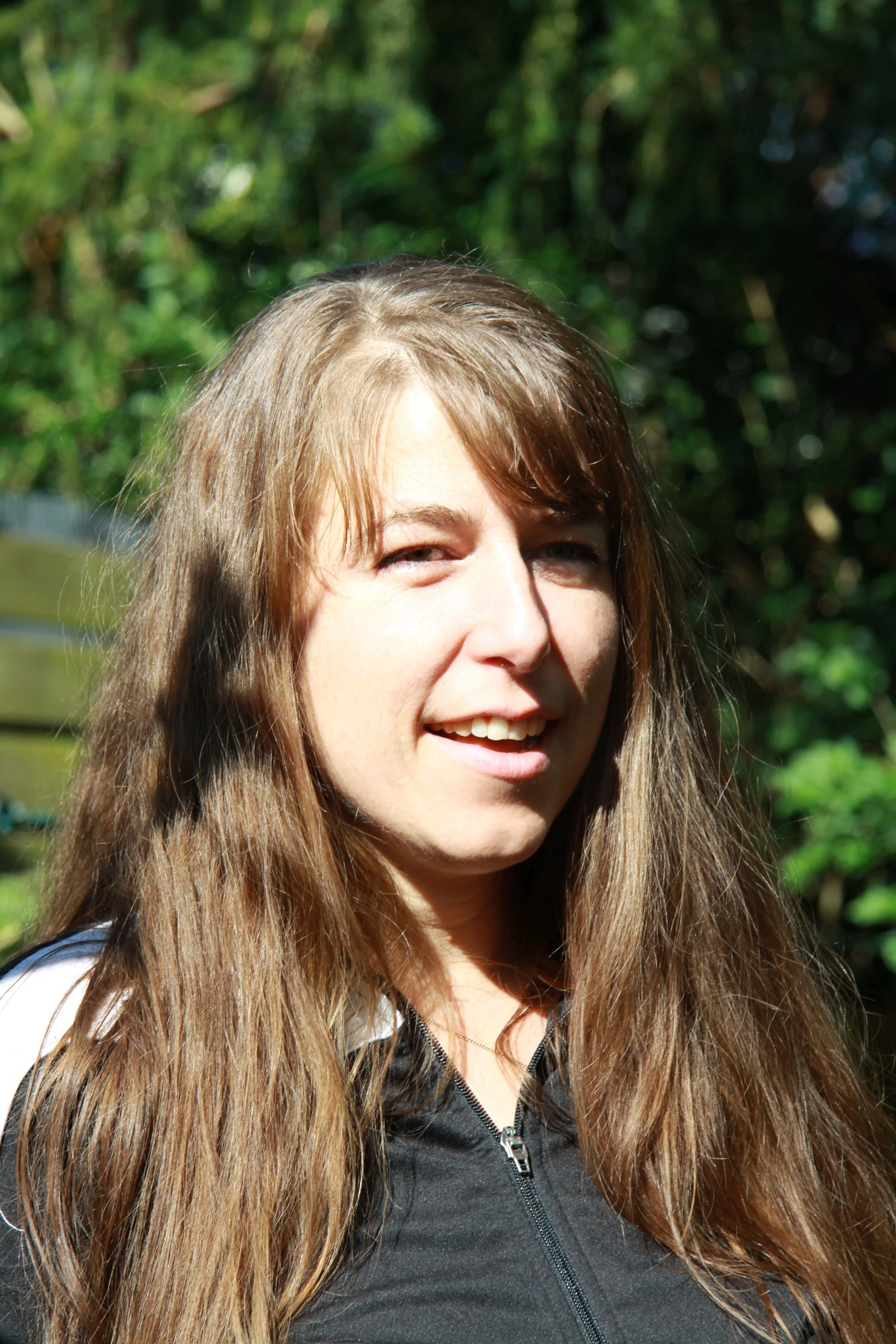 Lilian Gscheidel mit langen braunen Haaren im Sonnenlicht, lachend vor einem grünen, bewaldeten Hintergrund.