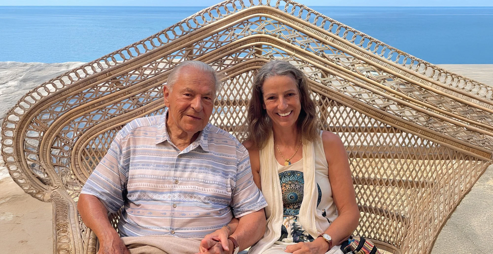 Brigitte Grof. Stan Grof. Ein älterer Mann und eine jüngere Frau sitzen gemeinsam in einem Rattan-Sofa am Strand, im Hintergrund das Meer und der Himmel.