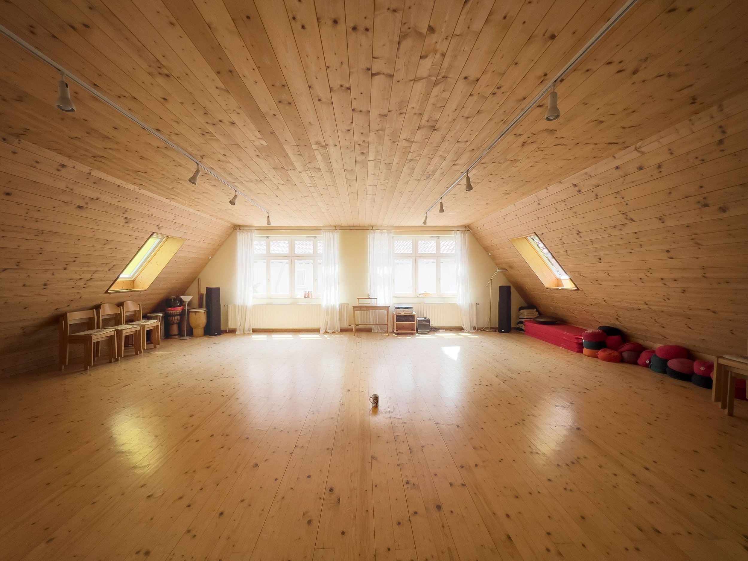 Ein heller Dachboden mit Holzvertäfelung, mehreren Fenstern, einem Lautsprecher und Yoga- oder Meditationskissen auf der rechten Seite.