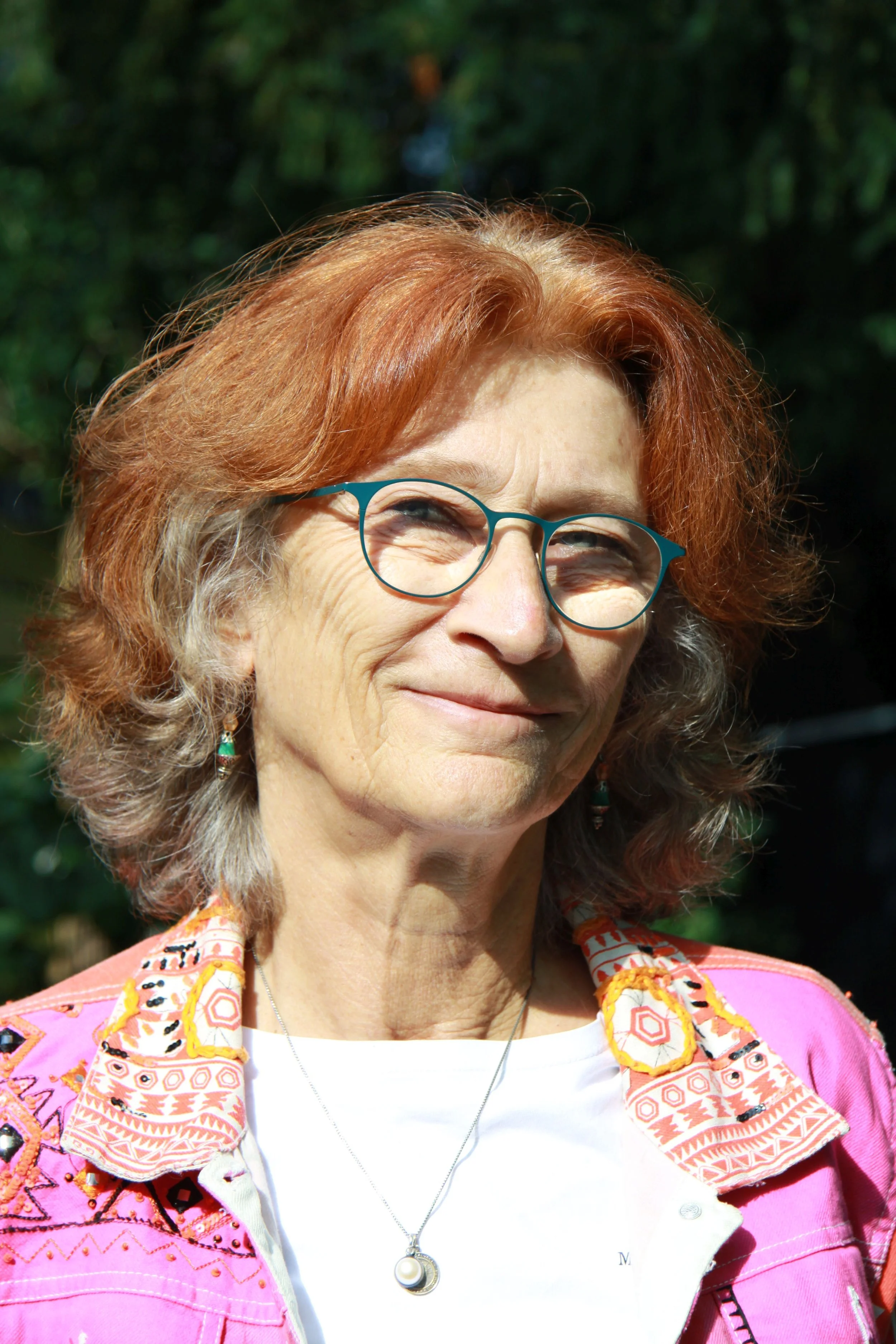 Kati Wortelkamp mit roten, lockigen Haaren und Brille, die in der Natur steht und in die Kamera lächelt.