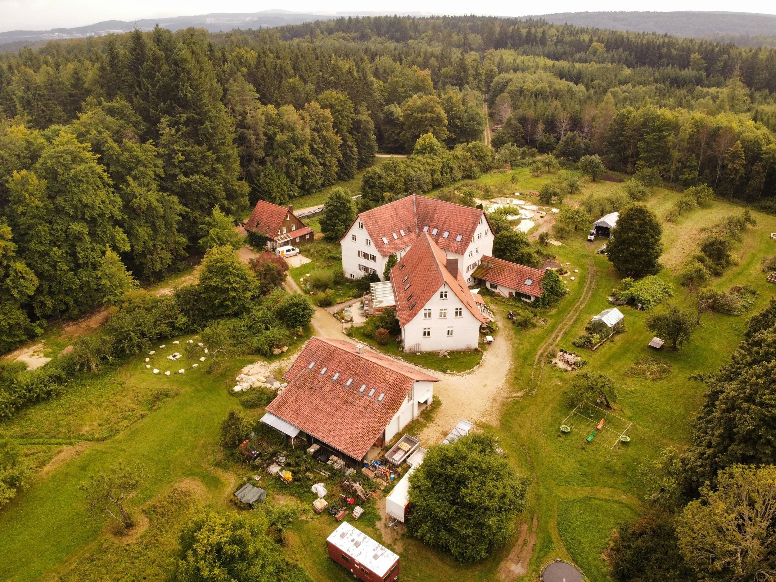 Luftaufnahme eines Hauses mit Rotziegel-Dach, umgeben von grünem Garten, Bäumen und Wiesen, vor einer dichten Waldlandschaft im Hintergrund.