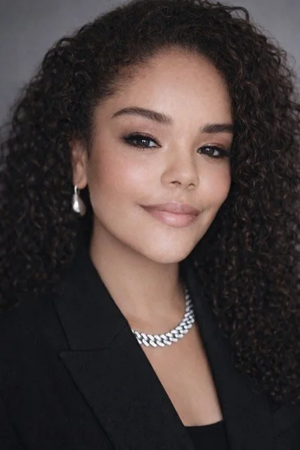 Young woman with curly hair wearing pearl earrings, a silver chain necklace, and a black blazer, smiling softly against a neutral background.